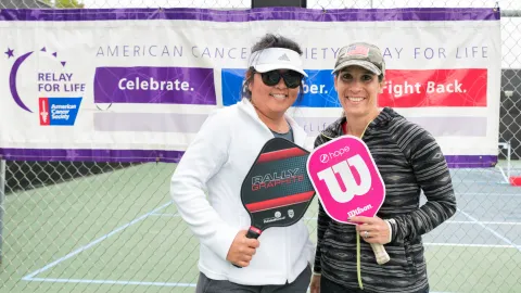 Two women holding pickleball paddles in front of American Cancer Society banner
